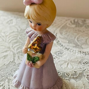 Puede incluir: Una figurita de porcelana de una ni&ntilde;a peque&ntilde;a con un vestido lavanda y sosteniendo un n&uacute;mero cuatro dorado. Tiene el pelo rubio con un lazo rosa y ojos azules.