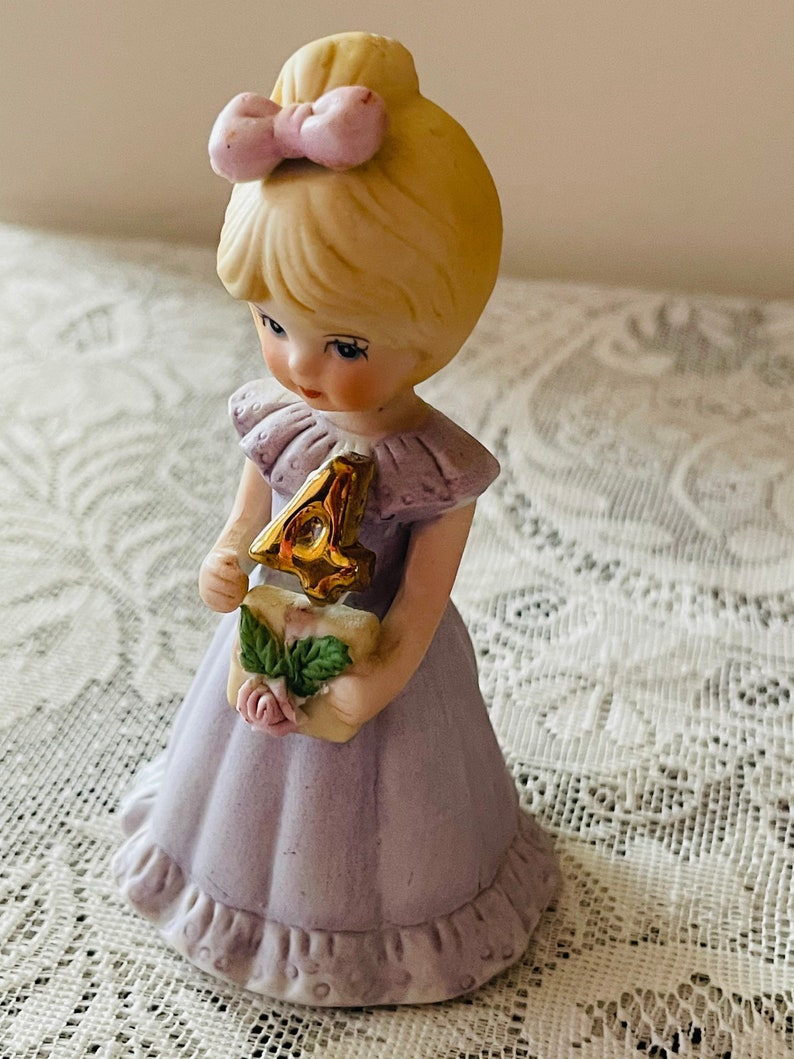 Puede incluir: Una figurita de porcelana de una ni&ntilde;a peque&ntilde;a con un vestido lavanda y sosteniendo un n&uacute;mero cuatro dorado. Tiene el pelo rubio con un lazo rosa y ojos azules.