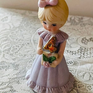 Puede incluir: Una figurita de porcelana de una ni&ntilde;a con un vestido lavanda que sostiene un n&uacute;mero cuatro dorado. Tiene el pelo rubio con un lazo rosa y una expresi&oacute;n dulce.