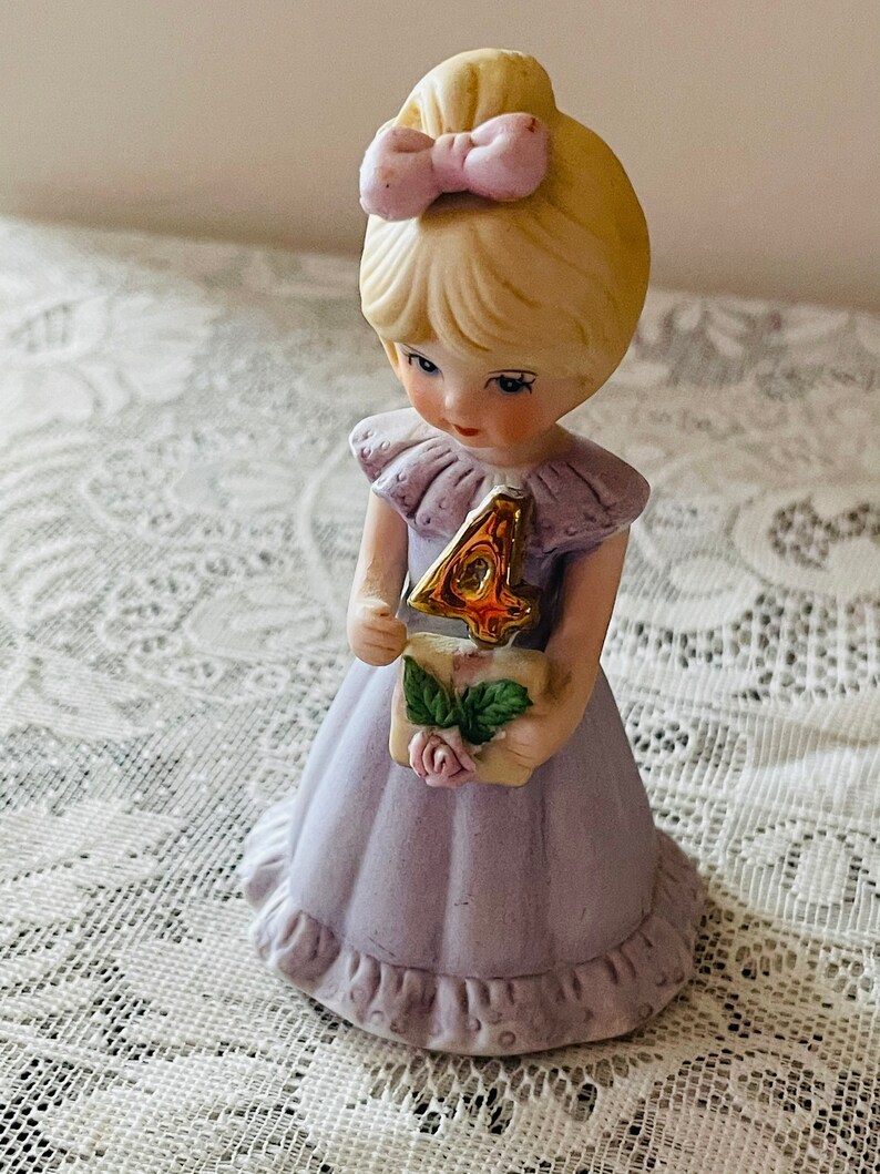 Puede incluir: Una figurita de porcelana de una ni&ntilde;a con un vestido lavanda que sostiene un n&uacute;mero cuatro dorado. Tiene el pelo rubio con un lazo rosa y una expresi&oacute;n dulce.