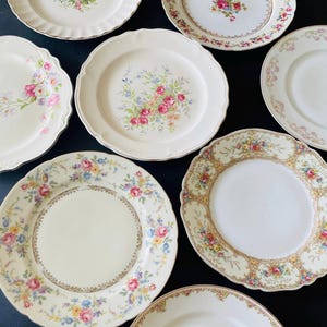 May include: Set of eight vintage floral patterned ceramic plates. The plates are white with a variety of floral designs in shades of pink, yellow, and blue. The plates have a gold trim around the edges.