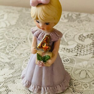 Puede incluir: Una figurita de porcelana de una ni&ntilde;a con un vestido lavanda que sostiene un n&uacute;mero cuatro dorado. Tiene el pelo rubio con un lazo rosa y lleva un cuello de volantes rosa y blanco.