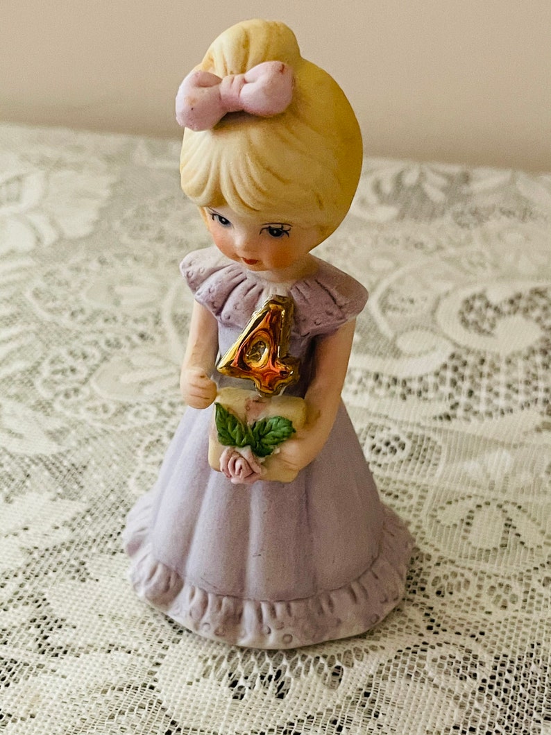 Puede incluir: Una figurita de porcelana de una ni&ntilde;a con un vestido lavanda que sostiene un n&uacute;mero cuatro dorado. Tiene el pelo rubio con un lazo rosa y lleva un cuello de volantes rosa y blanco.