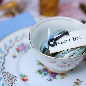 A mismatched vintage china teacup  and saucer stacked on top of a vintage china plate with a skeleton key and ribbon inside with a place card that says Tweedle Dee. All placed on table at a Alice in Wonderland Party