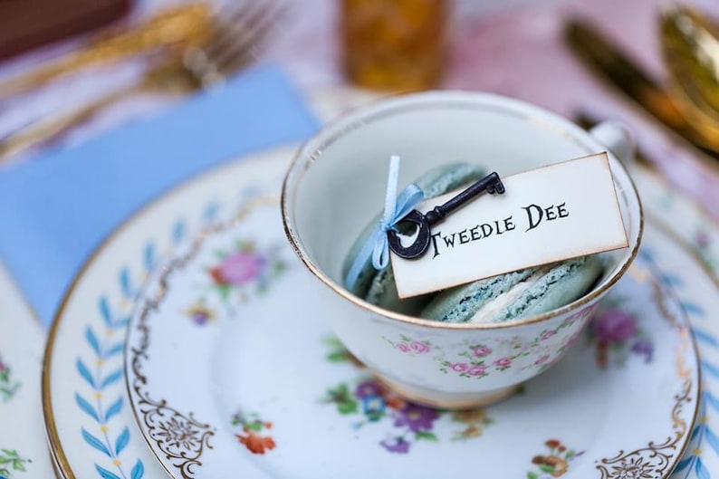 A mismatched vintage china teacup  and saucer stacked on top of a vintage china plate with a skeleton key and ribbon inside with a place card that says Tweedle Dee. All placed on table at a Alice in Wonderland Party