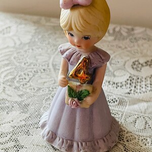 Puede incluir: Una figurita de porcelana de una ni&ntilde;a peque&ntilde;a con un vestido lavanda y sosteniendo un n&uacute;mero cuatro dorado. Tiene el pelo rubio con un lazo rosa y una expresi&oacute;n dulce.