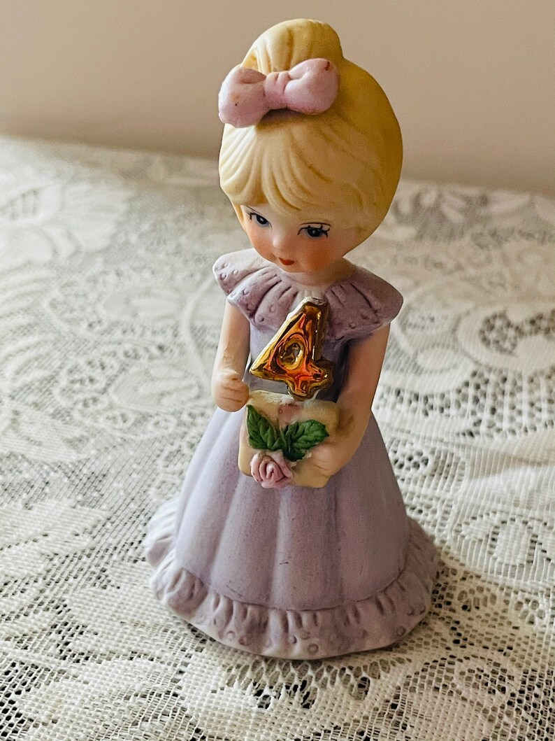 Puede incluir: Una figurita de porcelana de una ni&ntilde;a peque&ntilde;a con un vestido lavanda y sosteniendo un n&uacute;mero cuatro dorado. Tiene el pelo rubio con un lazo rosa y una expresi&oacute;n dulce.