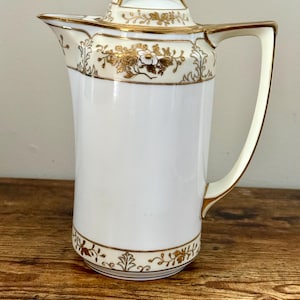 May include: A white porcelain pitcher with a lid, featuring gold floral and leaf patterns. The pitcher has a handle and a spout. The rim, handle, and lid are trimmed with gold. The pitcher is likely for serving beverages.