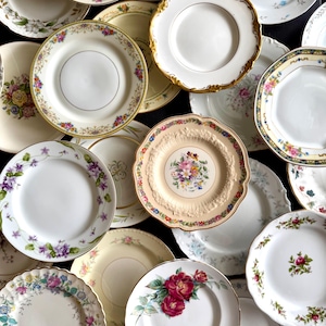May include: A collection of vintage porcelain plates with floral patterns. The plates feature various designs, including roses, wildflowers, and other colorful blooms. Some plates have gold rims, while others have scalloped edges. The plates are arranged on a dark surface.
