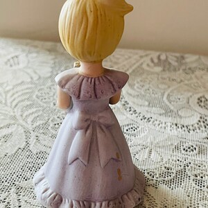 Puede incluir: Una figurita de porcelana de una ni&ntilde;a peque&ntilde;a que lleva un vestido lavanda con un borde de encaje blanco y un lazo. Tiene el pelo rubio y sostiene un peque&ntilde;o ramo de flores.