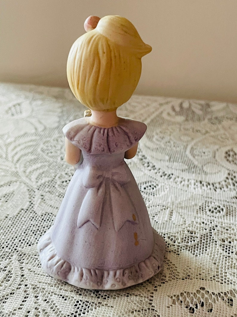 Puede incluir: Una figurita de porcelana de una ni&ntilde;a peque&ntilde;a que lleva un vestido lavanda con un borde de encaje blanco y un lazo. Tiene el pelo rubio y sostiene un peque&ntilde;o ramo de flores.