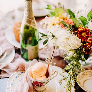 A vintage china teacup with a floral design filled with peach-colored ice cream. A silver spoon is in the teacup. The teacup is on a white plate with a floral design. A pink sheer cloth napkin is on the left side of the plate. A bottle of champagne is in the background.