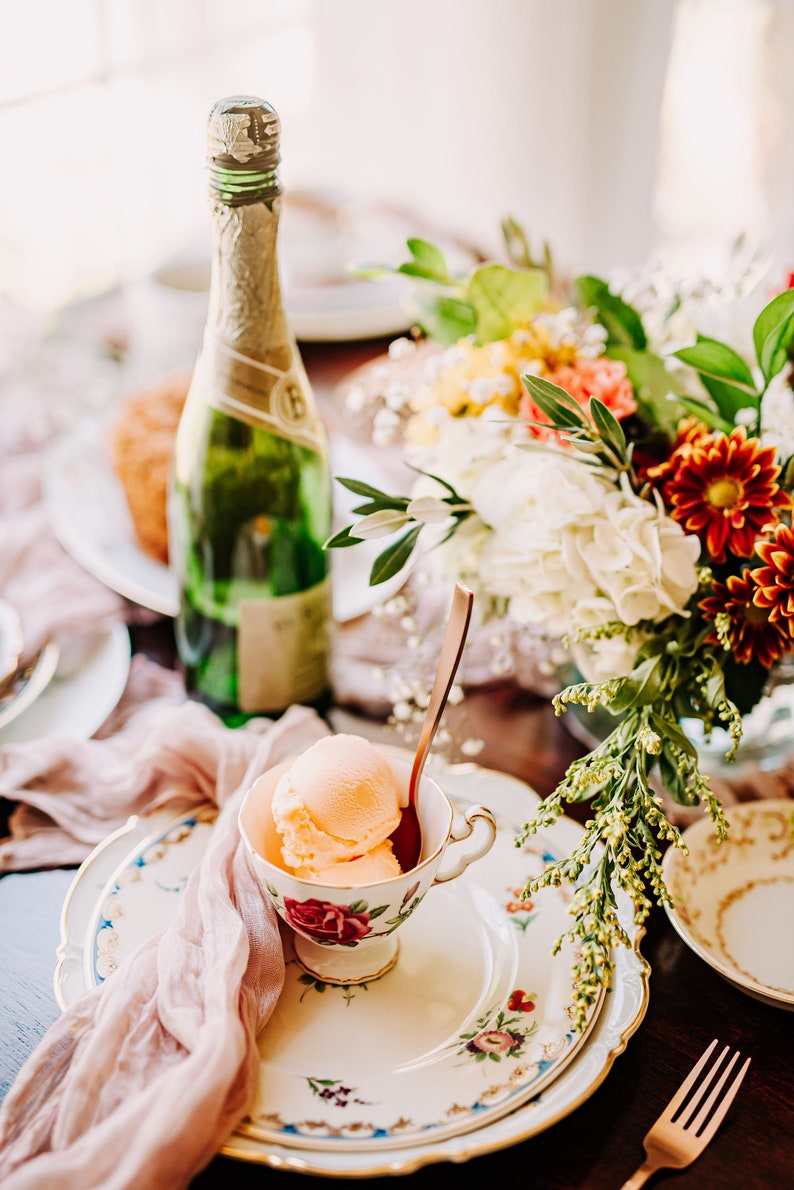 A vintage china teacup with a floral design filled with peach-colored ice cream. A silver spoon is in the teacup. The teacup is on a white plate with a floral design. A pink sheer cloth napkin is on the left side of the plate. A bottle of champagne is in the background.