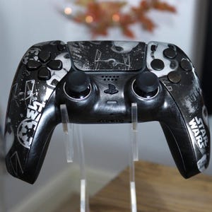 May include: A black and white wireless video game controller featuring a Star Wars theme. The surface displays graphic illustrations of TIE fighters, the Death Star, and the Imperial logo, with the Star Wars logo printed on the right handle.