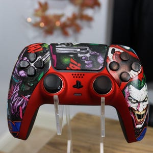 May include: A red video game controller featuring a custom graphic wrap with comic book style illustrations of a character with green hair and a wide grin, alongside a handgun graphic on the central touchpad and various colourful abstract patterns.