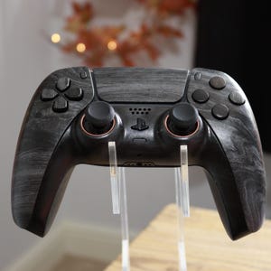 May include: A black video game controller featuring a dark grey wood grain texture on the front shell. The device includes two black analogue sticks with copper-coloured rings at the base, a central PlayStation logo, and standard directional buttons.