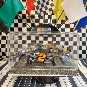 May include: A NASCAR diecast model car in a clear display case. The car is black, blue, and yellow with the number 2 on the side. The display case sits on a black base, with checkered flags in the background.