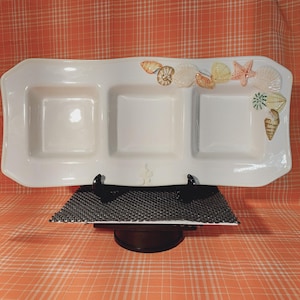 May include: A white ceramic serving dish with three compartments, decorated with seashell motifs. The dish is rectangular with a wavy edge and sits on a black stand. The seashells are in shades of beige, orange, and pink.