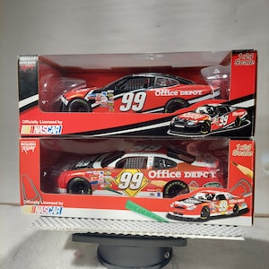 May include: Two unopened boxes of 1:24 scale NASCAR diecast cars. The boxes are red, black, and white with the Roush Racing logo. The cars are red, black, and white with the number 99 and "Office Depot" printed on the side. The boxes are on a gray surface.