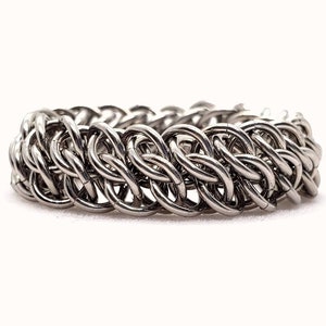 以下が含まれることがあります： 白い背景に、金属製のリングが互いに絡み合った銀色のチェーンメイルリングが置かれています。