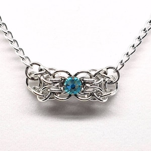 Puede incluir: Un collar de cadena de plata con un colgante de cristal azul. El colgante es un diseño tejido con un solo cristal en el centro.