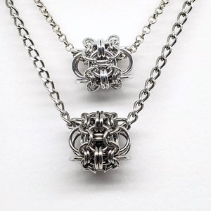 May include: A silver chain necklace with two silver metal chainmaille pendants. The larger pendant is a sphere and the smaller pendant is a cube.