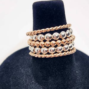Puede incluir: Un conjunto de cinco anillos apilables con cuentas alternas de plata y oro rosa. Los anillos son de metal y tienen un acabado pulido.