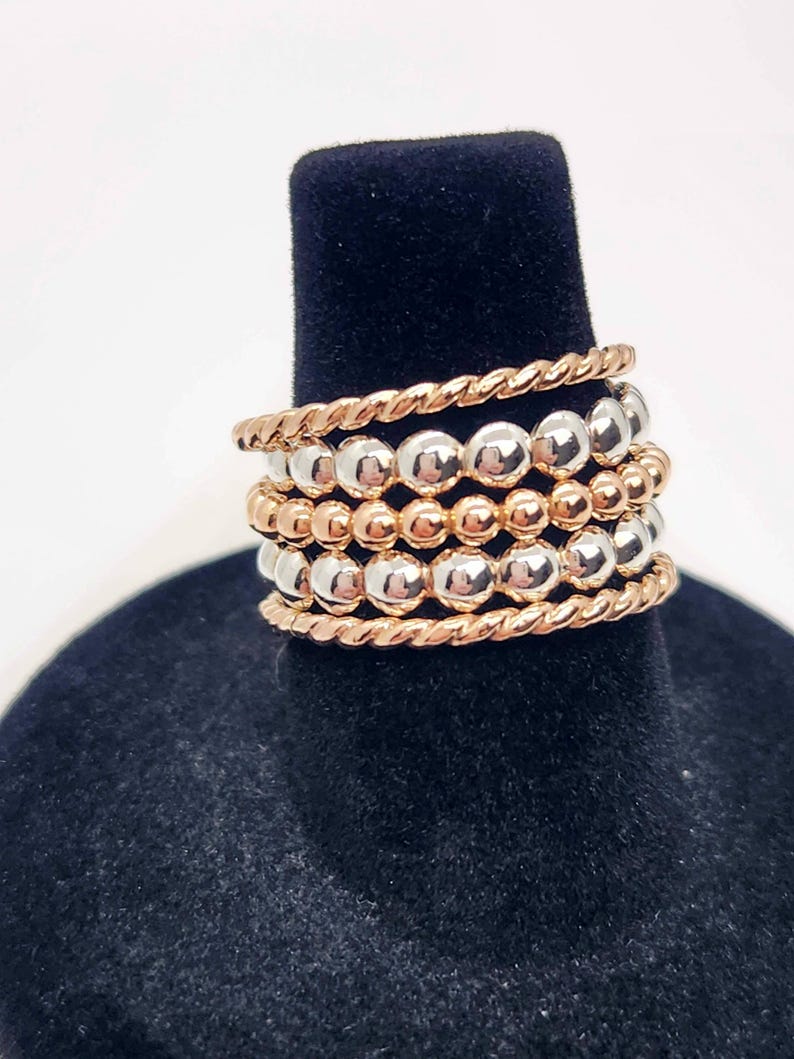 Puede incluir: Un conjunto de cinco anillos apilables con cuentas alternas de plata y oro rosa. Los anillos son de metal y tienen un acabado pulido.