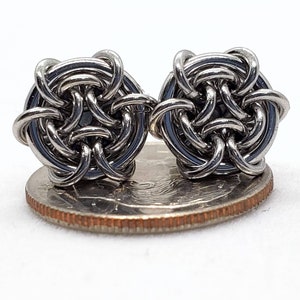 May include: A pair of silver-toned metal earrings with a woven knot design. The earrings are shown next to a UK 20 pence coin for size comparison.