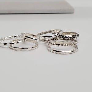 Puede incluir: Un conjunto de cinco anillos de plata, cada uno con un dise&ntilde;o diferente. Los anillos est&aacute;n apilados uno encima del otro, con el anillo m&aacute;s adornado en la parte superior.