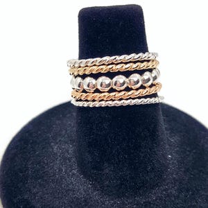 Puede incluir: Un conjunto de cinco anillos apilables de plata y oro. Los anillos est&aacute;n hechos de diferentes texturas, incluyendo lisos, con cuentas y retorcidos. Los anillos est&aacute;n apilados en un expositor de anillos negro.