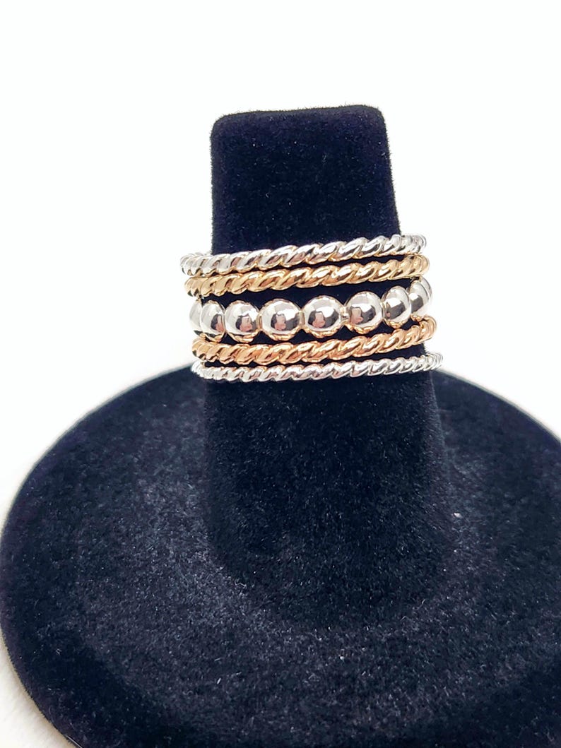 Puede incluir: Un conjunto de cinco anillos apilables de plata y oro. Los anillos est&aacute;n hechos de diferentes texturas, incluyendo lisos, con cuentas y retorcidos. Los anillos est&aacute;n apilados en un expositor de anillos negro.