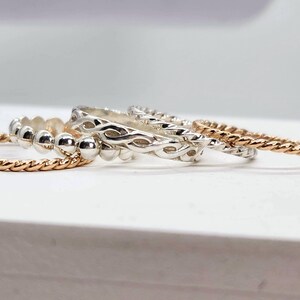 Puede incluir: Cuatro anillos de plata apilados uno encima del otro. El anillo superior tiene un dise&ntilde;o trenzado, el segundo anillo tiene un dise&ntilde;o de cuentas, el tercer anillo tiene un dise&ntilde;o trenzado y el anillo inferior es una banda delgada y retorcida.