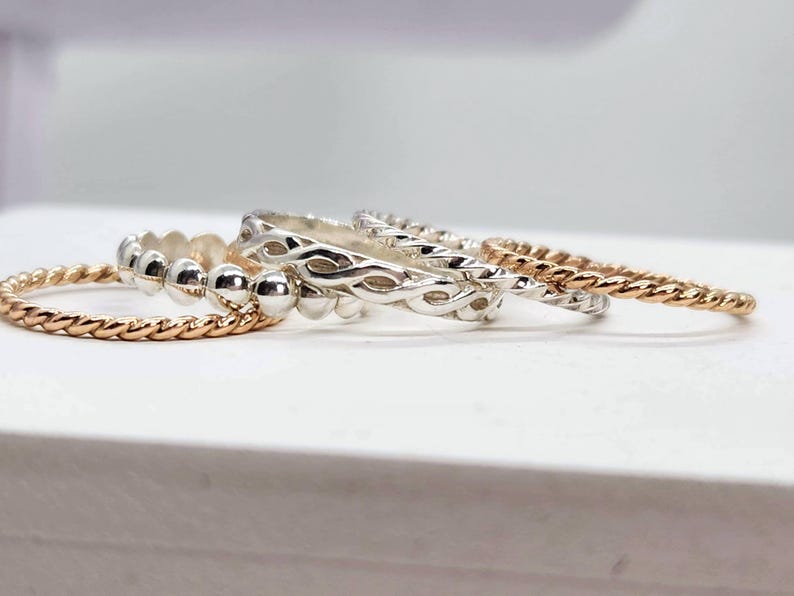 Puede incluir: Cuatro anillos de plata apilados uno encima del otro. El anillo superior tiene un dise&ntilde;o trenzado, el segundo anillo tiene un dise&ntilde;o de cuentas, el tercer anillo tiene un dise&ntilde;o trenzado y el anillo inferior es una banda delgada y retorcida.