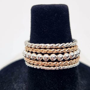 Puede incluir: Un conjunto de cinco anillos apilables de plata y oro. Los anillos est&aacute;n hechos de alambre trenzado y tienen una fila de peque&ntilde;as cuentas de plata.