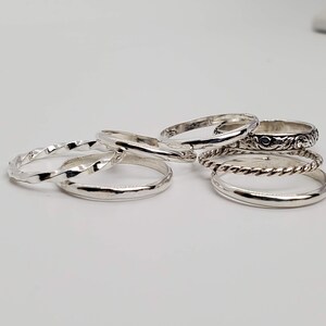 Puede incluir: Un conjunto de seis anillos de plata con diferentes texturas y dise&ntilde;os. Los anillos est&aacute;n apilados uno encima del otro.
