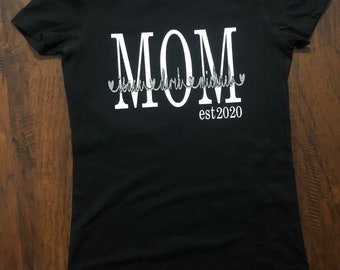 May include: Black t-shirt with white text that reads "MOM est. 2020" with the word "MOM" in large letters and the rest of the text in a smaller font. The text is outlined in silver glitter.