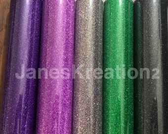 May include: Five rolls of glitter vinyl in different colors: purple, pink, silver, green, and black. The rolls are lined up side by side on a wooden surface.