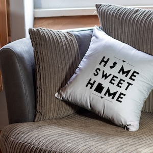 May include: White throw pillow with black text that reads "Home Sweet Home" with a small outline of the state of New Jersey in place of the "O" in "Home". The pillow is on a gray couch with a brown corduroy cover.