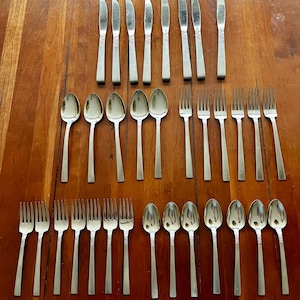 Peut inclure: Un ensemble de couverts en acier inoxydable, comprenant des couteaux, des cuillères et des fourchettes, disposés sur une surface en bois. Les couverts présentent un design simple et élégant, reflétant la lumière. L'arrangement suggère un cadre de restauration formel.