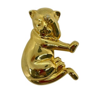 May include: A gold-toned metal brooch in the shape of a cat sitting with its head turned to the side. The cat's eyes are closed and its mouth is slightly open.