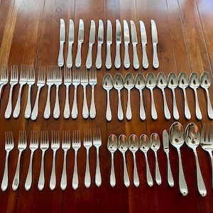 May include: A full set of stainless steel cutlery, including forks, knives, and spoons, arranged on a wooden surface. The cutlery has a polished finish, reflecting the light. The set includes various sizes of forks and spoons.