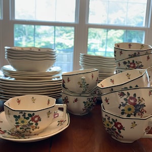 Puede incluir: Un juego de vajilla de porcelana blanca con un patrón floral en tonos rojos, verdes y amarillos. El juego incluye platos, cuencos y tazas de té con platillos a juego.