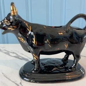 May include: Black ceramic creamer in the shape of a cow with gold accents. The cow is standing on a black base.