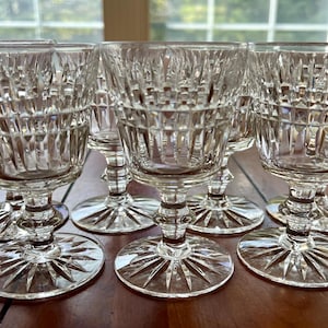 Może przedstawiać: Sześć przezroczystych szklanek na nóżce z grawerowanym wzorem. Szklanki są ułożone w półkole na drewnianej powierzchni.