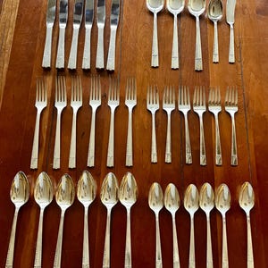 May include: A collection of silver-colored flatware, including knives, forks, spoons, and butter knives, arranged on a wooden surface. The silverware has a decorative pattern on the handles. The arrangement suggests a formal dining setting.