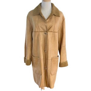 May include: A tan-colored coat with a shearling collar and cuffs. The coat has a button-up front, two front pockets, and a longline silhouette. The coat is displayed on a mannequin. The coat is made of a suede-like material.