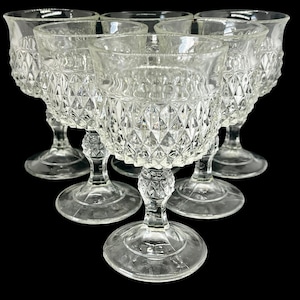 May include: A set of six clear, vintage-style goblets. Each glass features a diamond-cut pattern on the bowl and a decorative stem. The glasses are arranged in a semi-circular formation against a black background, showcasing their intricate design.