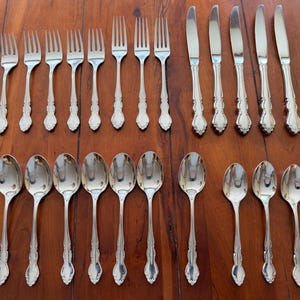 May include: A collection of silver-colored flatware, including forks, knives, and spoons, arranged on a wooden surface. The silverware has decorative handles and a polished finish, reflecting the light. The set includes multiple pieces of each utensil.