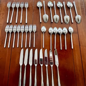 Pode incluir: Um conjunto de talheres de aço inoxidável, incluindo garfos, colheres e facas, dispostos sobre uma superfície de madeira. Os talheres têm um design simples e elegante, com cabos longos e finos e superfícies polidas. O conjunto inclui 12 garfos, 12 colheres e 12 facas.